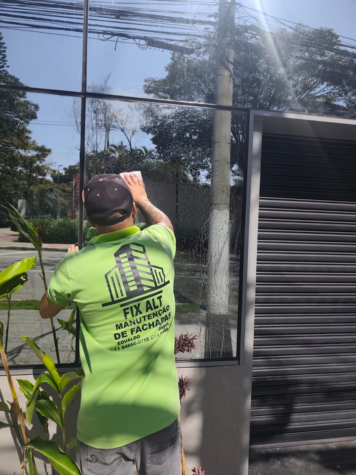 homem de costas com camisa verde limão limpando vidro de fachada de loja, com logo Fix Alt Manutenção de Facha