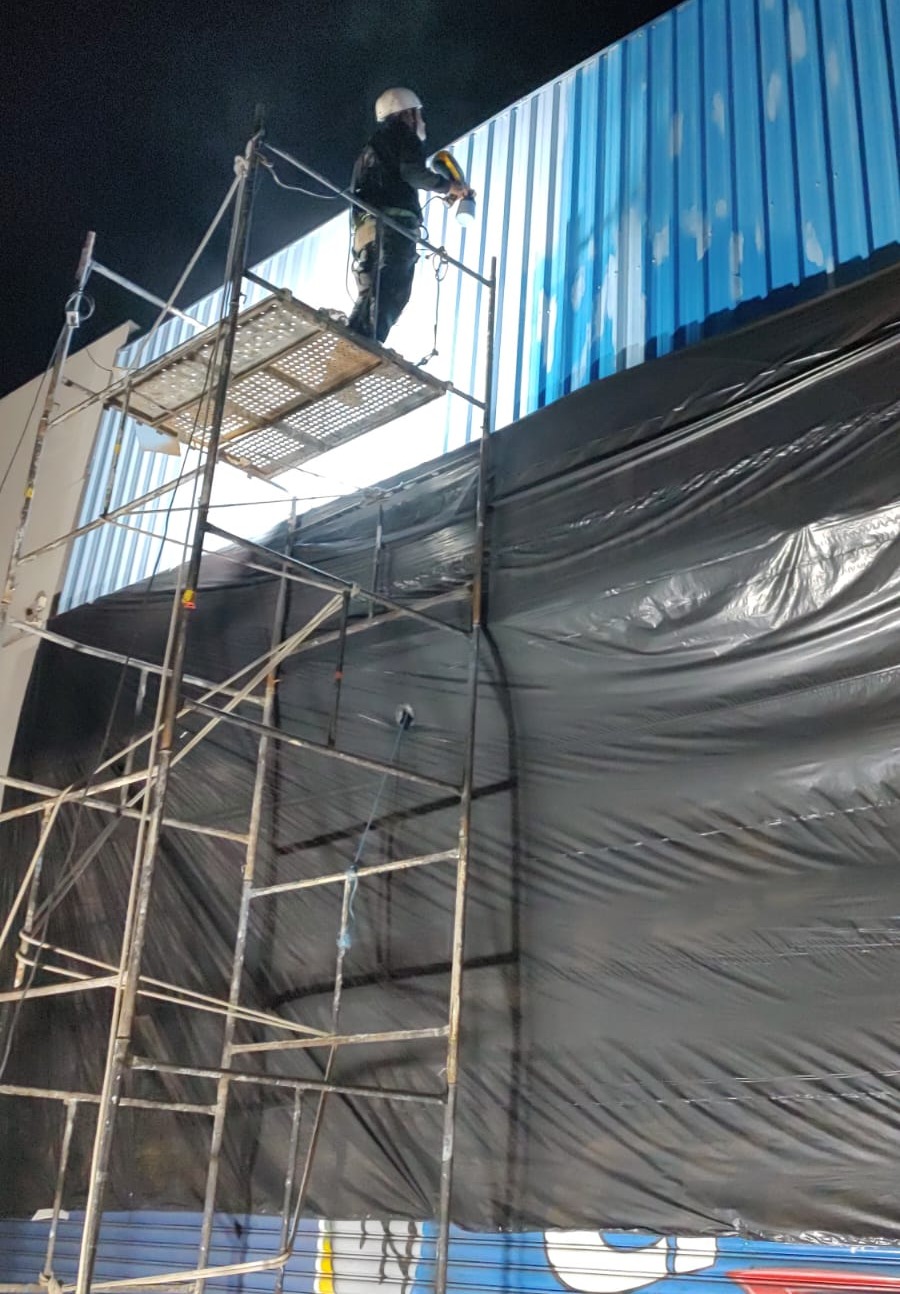 trabalhador de capacete sobre andaime pintando com pistola a fachada de telhado metálico azul à noite, com lon