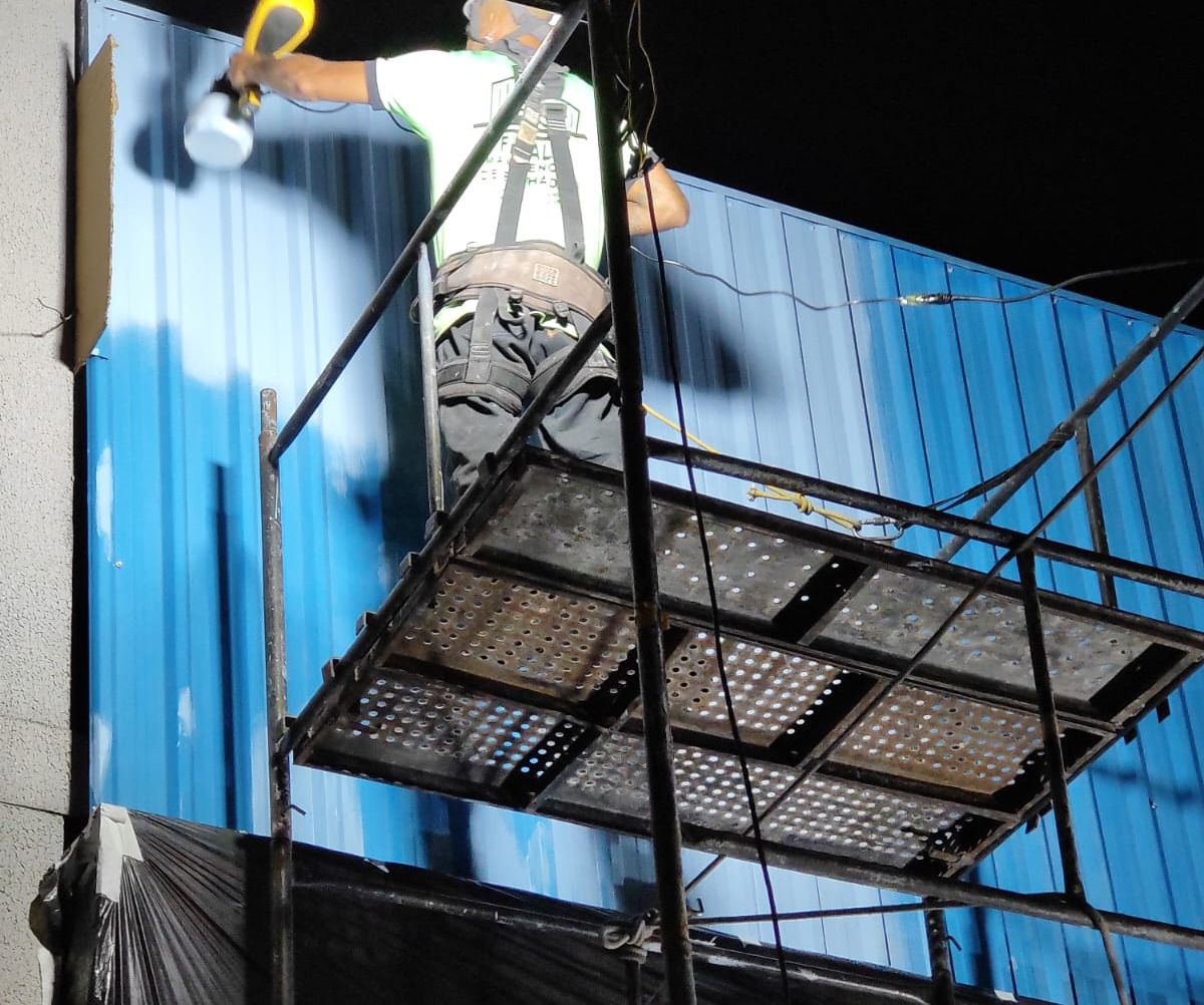 trabalhador com capacete e cinto de segurança sobre um andaime pintando com pistola uma fachada metálica azul