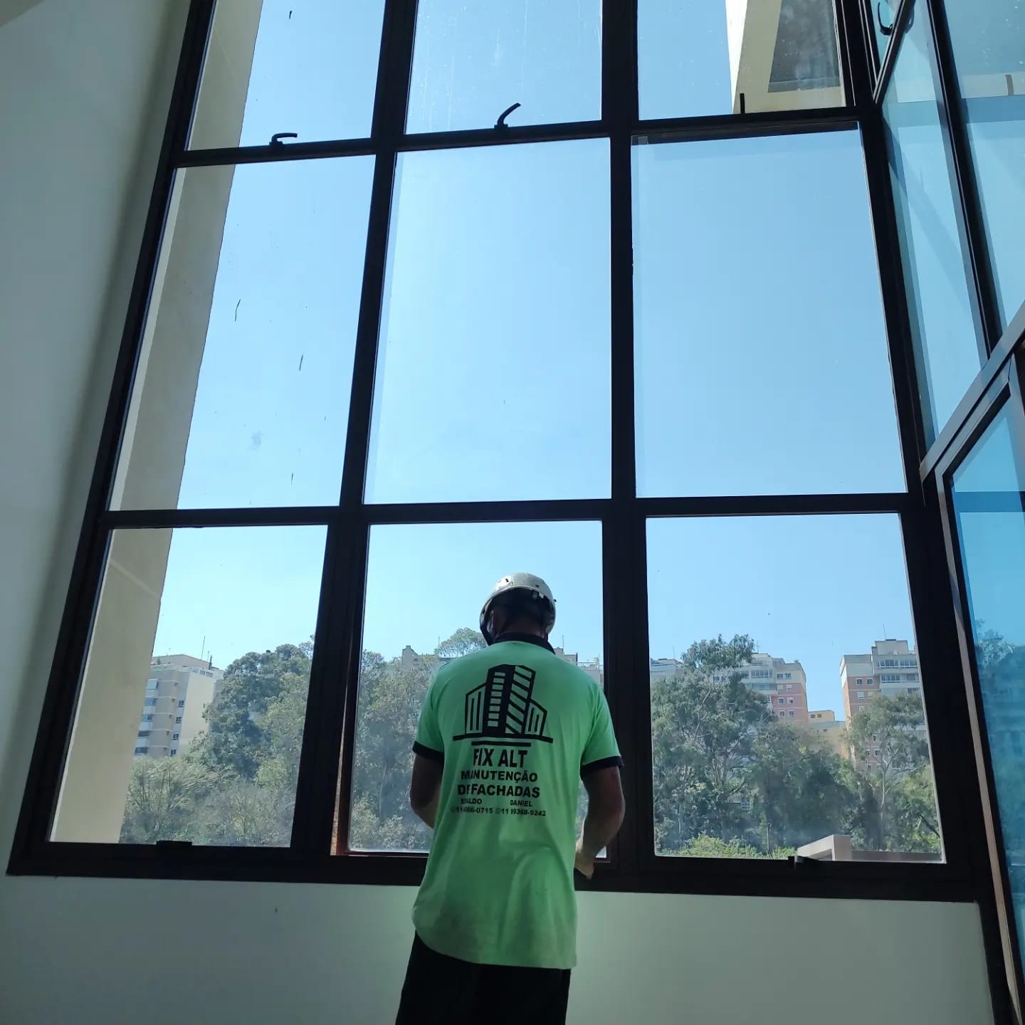 trabalhador de camiseta verde e capacete limpando/mantendo grandes janelas de vidro de uma fachada, com prédio