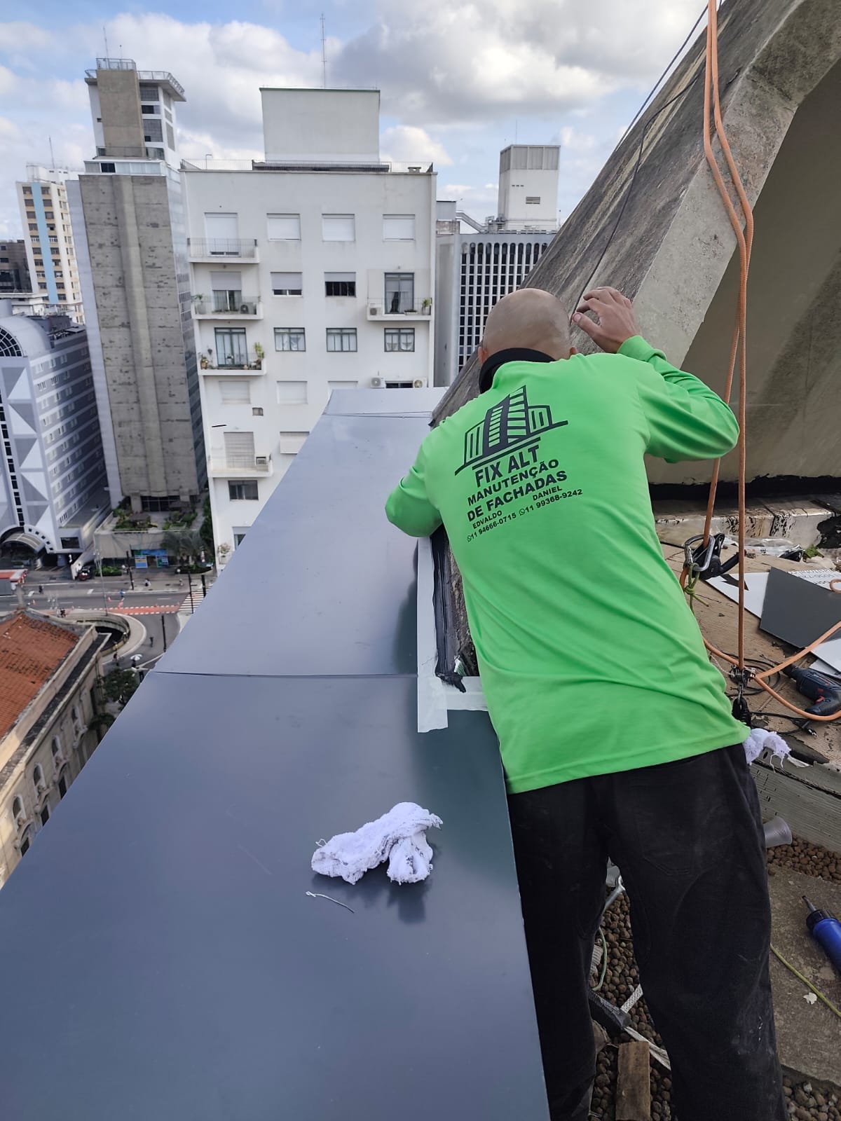 técnico de camisa verde com logo Fix Alt trabalhando em cobertura de fachada na altura, com prédios da cidade
